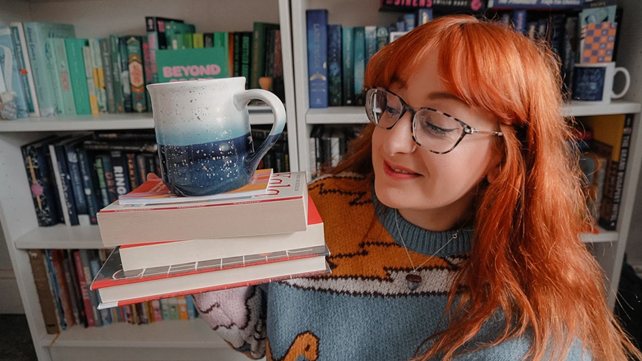 Ginger girl holding a stack of books with a mug balancing on top, sitting in front of her bookshelves.
